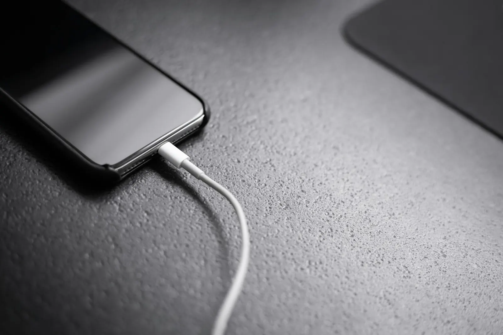 Schwarzes Smartphone mit eingestecktem weißem Ladekabel auf einem grauen Tisch